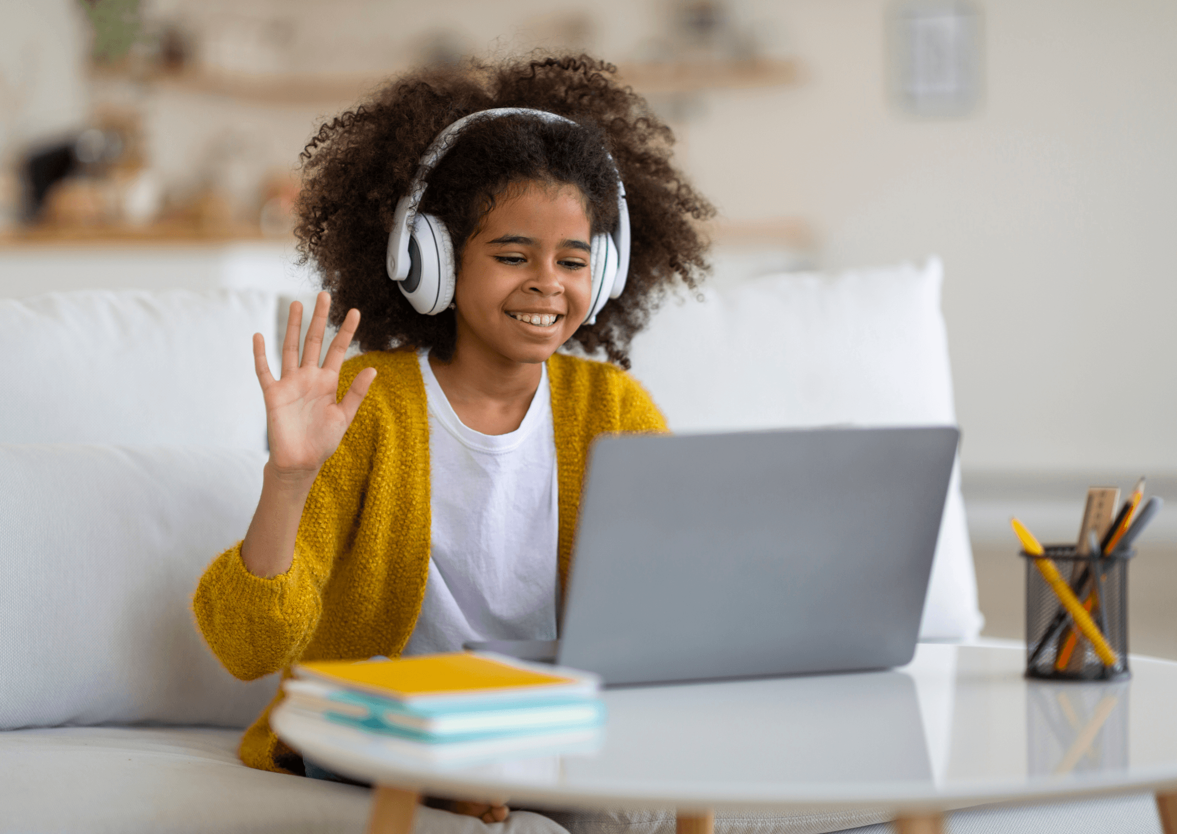 A student in headphones waving at her laptop, schoolbooks stacked on the table